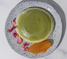 Load image into Gallery viewer, A round green dessert or cake with a pale cream border, served on a speckled gray plate with dried pink flowers and a dollop of orange sauce on a white marble surface. Nectar + Peel