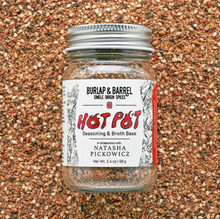Load image into Gallery viewer, A glass jar of Hot Pot Seasoning & Broth Base by Burlap & Barrel, filled with a brown spice blend, set against a background of loose ground spices. The label features red and black text with a white background and a red side stripe.