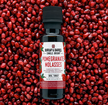 Load image into Gallery viewer, A bottle of Burlap & Barrel Pomegranate Molasses centered against a dense background of red pomegranate seeds. The bottle has a black cap and a white label with red and green text and illustrations.