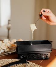 Load image into Gallery viewer, A black fondue pot on a wooden table, with a hand holding a fork lifting melted cheese above the pot. Sliced bread sits in a glass dish in the background.