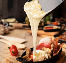 Load image into Gallery viewer, Melted cheese being poured from a pan into a cast-iron skillet filled with breakfast foods including sausage, bacon, mushrooms, bread, and cream, set on a wooden table with blurred kitchen items in the background.