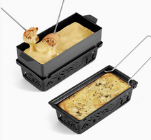 Load image into Gallery viewer, Two black cast-iron cheese raclette pans with decorative cutout sides and metal handles, shown on a white background. One pan contains melted yellow cheese with toasted bread on skewers, and the other contains a baked cheesy casserole with herbs.