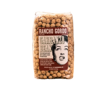 Load image into Gallery viewer, A clear plastic bag of Rancho Gordo Garbanzo Beans filled with light tan chickpeas, standing upright on a burlap cloth against a rustic brown background.