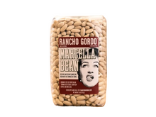 Load image into Gallery viewer, A clear plastic bag of Rancho Gordo Marcella Bean dried beans, standing upright on a piece of burlap fabric against a rustic wooden and burlap background.