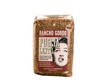 Load image into Gallery viewer, A bag of Rancho Gordo Puglia Lentil whole grain lentils, displayed on a rustic wooden surface with a burlap cloth underneath and a blurred burlap sack in the background.