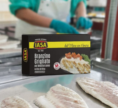 IASA Grilled Branzino in a tin can, in a black box with yellow stripe, next to hand-filleted fish at factory with person in background working.  Italy