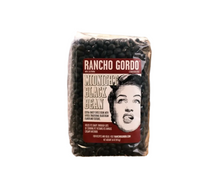 Load image into Gallery viewer, A bag of Rancho Gordo Midnight Black Bean coffee beans sits on a piece of burlap fabric on a wooden table, with a blurred burlap sack in the background.
