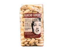 Load image into Gallery viewer, A bag of Rancho Gordo Royal Corona Bean dried beans standing upright on a burlap mat, with blurred burlap sacks in the background.
