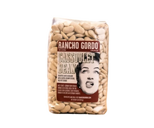 Load image into Gallery viewer, A bag of Rancho Gordo Cassulet Bean, made from green beans, standing on a piece of burlap over a rustic wooden surface with a blurred burlap background.
