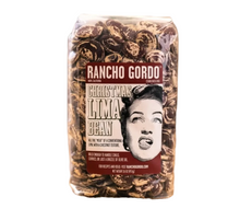 Load image into Gallery viewer, A clear plastic bag of Rancho Gordo Christmas Lima Bean filled with dark brown marbled beans, resting on a piece of burlap over a rustic wooden surface.
