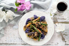 Load image into Gallery viewer, White plate of beautiful braised spring vegetables (purple cauliflower, green asparagus and brown mushrooms), on a white crackly wood table, alongside fork, glass of red wine, white and pink azaleas and grey linen towel.

